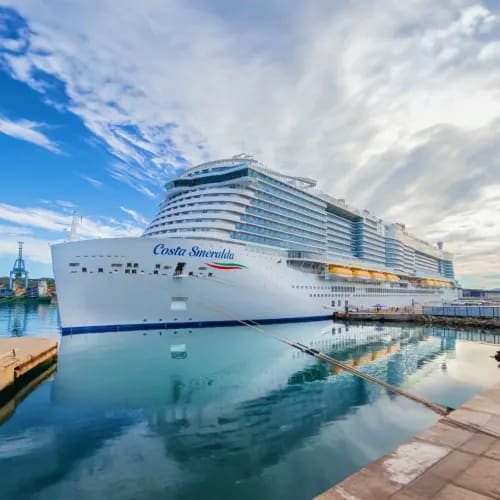 Large white Costa Smeralda cruise ship docked at port with blue water and cloudy sky, winter cityscape in background